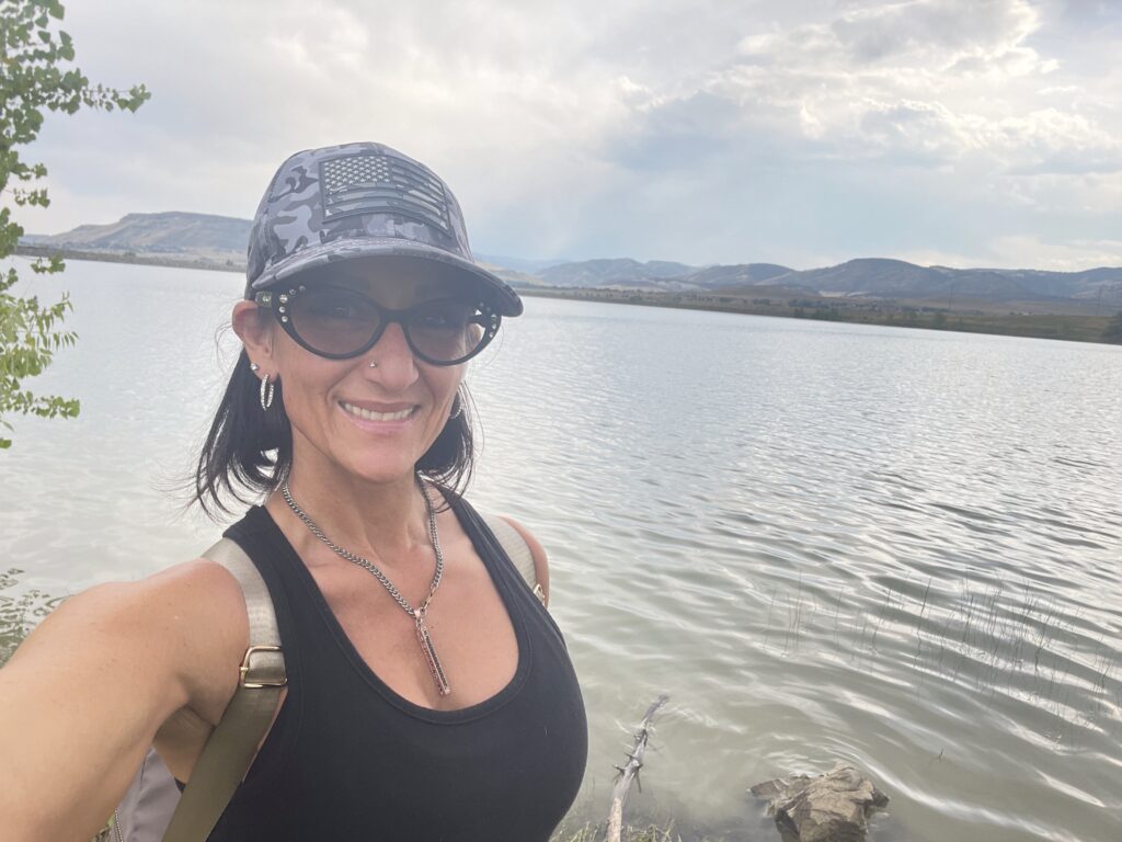 Anisa is ElderFIT's Personal Trainer for Seniors in Denver and she is standing in front of a lake on a summer day hiking and smiling at the camera.