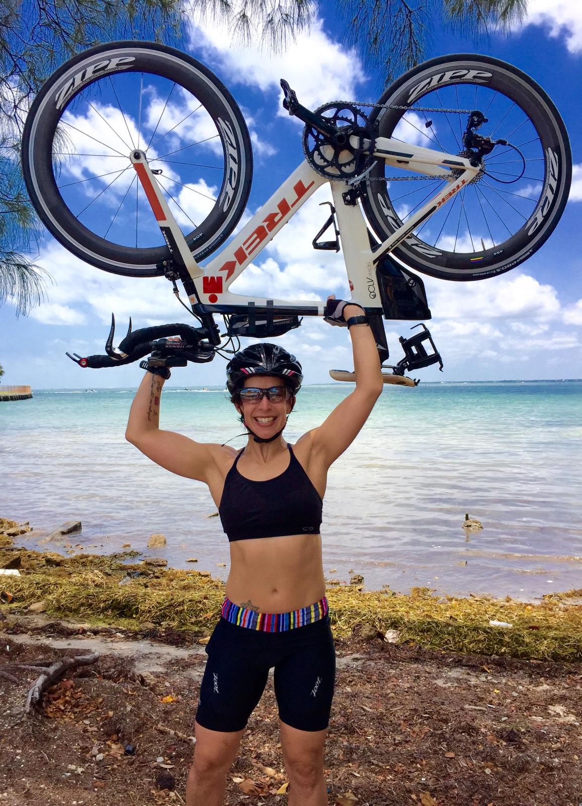 Personal trainer for seniors in Miami is holding her road cycling bike over her head, smiling, standing in front of a body of water and a beach.