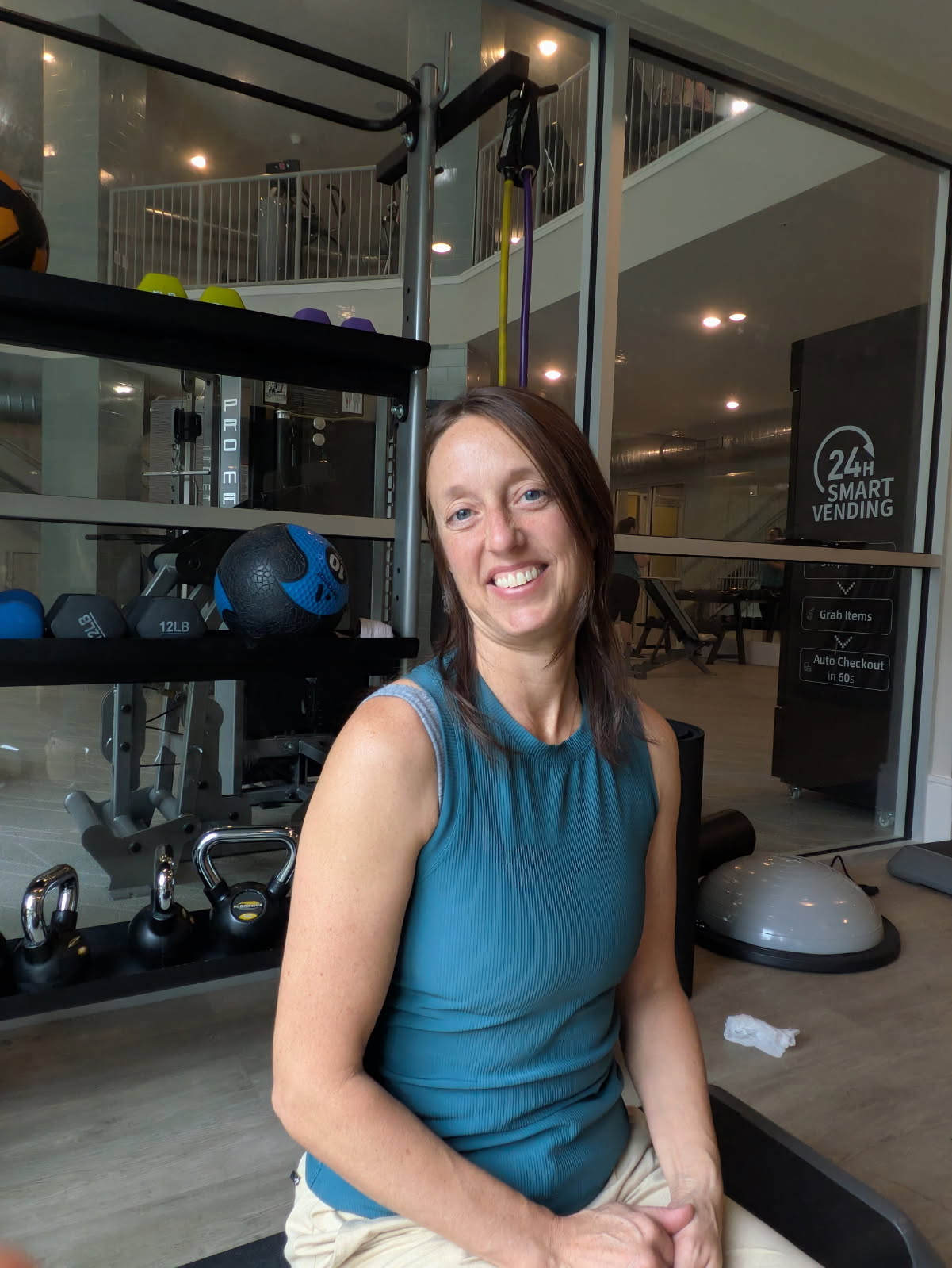 A pic of Claire, a top ElderFIT personal trainer for seniors in Jacksonville sits in a gym with a rack of gym equipment behind her and smiles at the camera.