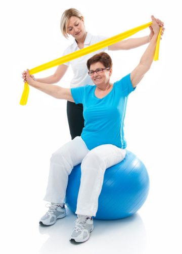 Senior aged female sitting on exercise ball with trainer behind